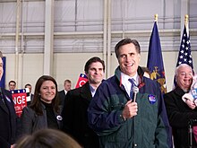 Mitt Romney surrounded by people, holding a microphone and smiling