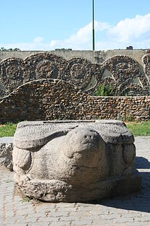 Photographie d'un dragon-tortue (bixi) de face, avec derrière des anciens murs avec des motifs indéterminés.