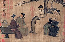 Four men dressed in robes and black square cut hats gather around a tree talking to one another. Three are sitting on rocks while the fourth is leaning over a horizontally bent branch of the tree.