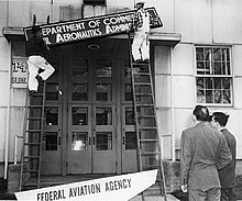 Рабочие на лестницах меняют старую вывеску «Управление гражданской авиации» на новую вывеску «Федеральное авиационное агентство» над высоким входом в здание.
