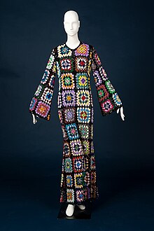 White mannequin on dark blue backdrop featuring a full-length jumpsuit with rainbow-colored sequins in clusters, mimicking the texture and composition of crochet granny squares