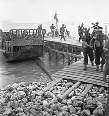 Soldiers disembark a landing craft at a makeshift wooden jetty