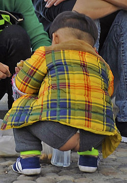 A very young boy, squatting and facing away from the camera, wearing a bright yellow plaid shirt and pants with an opening in the rear through which his buttocks show