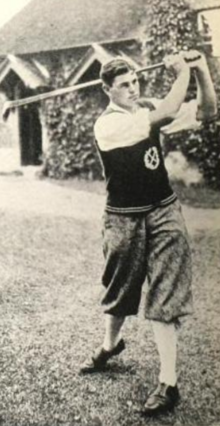 Un giovane uomo bianco in una posa da golf swing, che indossa pantaloni corti e un maglione lavorato a maglia