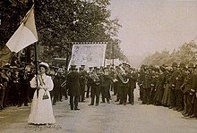 Dorothy Radcliffe holding aloft a purple, white and green flag in front of one of the seven bands Dorothy Radcliffe holding aloft a purple, white and green flag.jpg