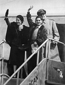 The MacArthur family standing at the top of the stairs leading from a passenger aircraft. Douglas MacArthur stands behind while his wife Jean and son Arthur wave to those below.