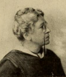 An older white woman with white hair in an updo, wearing pince-nez glasses on a lanyard, and a dark dress or academic robe, photographed in profile