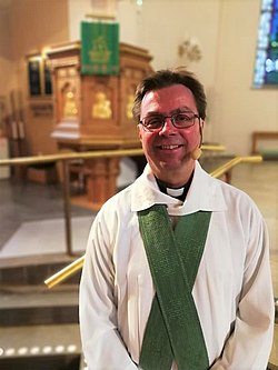 Göran Bondesson, leende. Klädd i vit alba och grön stola. I bakgrunden ambon i Sankt Johannes kyrka, Norrköping.