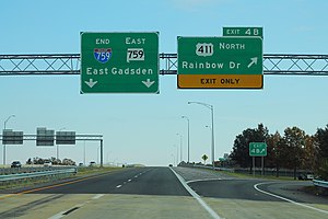 Two green signs are located above an elevated portion of roadway with no traffic visible on a cloudy day.