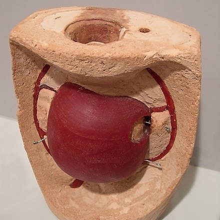 The hollow paraffin apple is covered with a final, fire-proof mould, in this case clay-based, an open view. The core is also filled with fire-proof material. Note the stainless steel core supports. In the next step (not shown), the mould is heated in an oven upside-down and the wax is "lost"