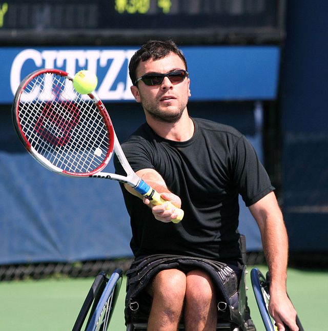 נועם גרשוני יושב על כיסא גלגלים במהלך משחק טניס. פניו לצופה, ידו השמאלי על גלגל הכיסא, וידו הימנית מניפה מחבט טניס לעבר כדור טניס האף לעברו.
