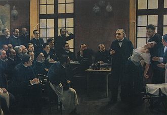 A painting of a 19th-century medical lecture. At the front of the class, a woman faints into the arms of a man standing behind her, as another woman, apparently a nurse, reaches to help. An older man, the professor, stands beside her and gestures as if making a point. Two dozen male students watch them.