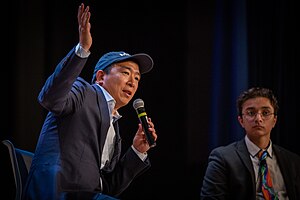 Yang is holding a microphone while gesturing and making a speech