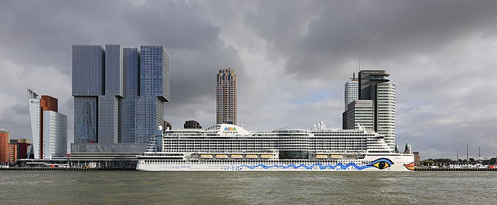 Ancorado em um cais um grande barco branco, o casco com uma linha azul.  Atrás de edifícios modernos, céu cinza.