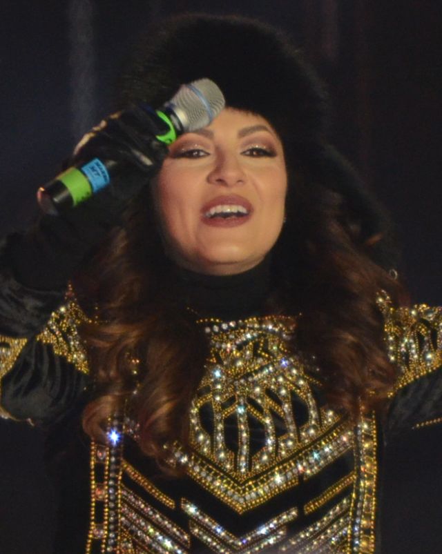 A woman is seen holding a microphone in her right hands; she wears a fur cap, black gloves and a gold-black outfit.