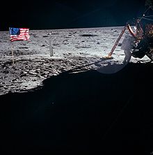 Un astronaute en combinaison se penche vers le module, un drapeau américain est à gauche.