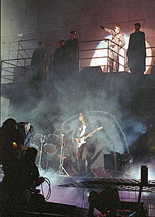 A concert stage in front of a wall with 2 levels. Five men stand on a balcony, including Roger Waters, who is saluting with his arm. On the lower level is a drum kit and a man playing guitar.