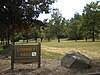 Im Vordergrund steht ein großer Felsen zusammen mit einem Schild mit dem Namen, der Verwaltung und den Öffnungszeiten des Parks;  im Hintergrund ist eine Wiese und vereinzelte Bäume.