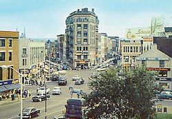 A view down on a busy city intersection with a tall wedge-shaped building at the junction of two streets that fork in the center. A tree and statue of a horse are at the bottom, in the foreground