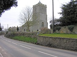 Stone building with square tower, separated from the road in the foreground by a stone wall.