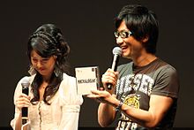 Hideo Kojima (with model Yumi Kikuchi) at the 2011 Tokyo Game Show holding an original Metal Gear Solid jewel case