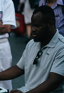 A black man in a grey polo shirt is seated at a table, facing left of frame.