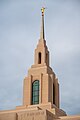 The spire of the Red Cliffs Utah Temple dominates the view of a portrait shot. A multilevel spire has rounded windows on the sides, along with a golden statue at the top of the the angel Moroni, against a backdrop of a blue sky.