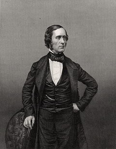 photograph of standing figure facing right, left hand on hip, right hand resting on chair