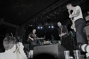 Swans tocando en vivo en Varsovia en diciembre de 2010. De izquierda a derecha: Christoph Hahn en lap steel, Michael Gira en guitarra, Thor Harris en percusión, Chris Pravdica en bajo y Norman Westberg en guitarra.  Phil Puleo está sentado detrás de la batería.