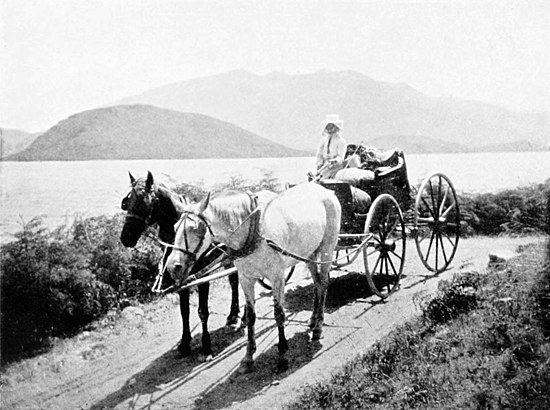 A horse-drawn carriage ("The Berline") with two horses pulling it and a driver. The carriage is on a dirt road with a lake and hills to the left.