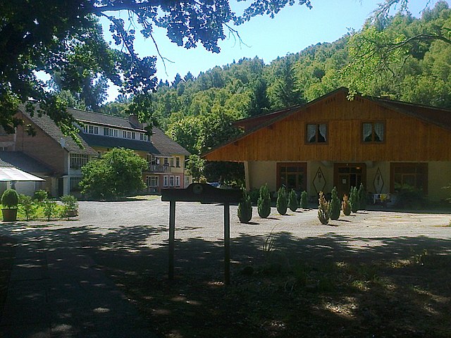 Das Foto zeigt den teilweise durch Bäume beschatteten Hof eines Anwesens bei sonnigen Wetter. Zwei Gebäude sind zu sehen, rechts eines im Chaletstil mit Erdgeschoss und ausgebautem Dachgeschoss, links ein größeres mit zwei Geschossen zuzüglich ausgebautem Dachgeschoss unter einem Satteldach. Hinter den Häusern steigt ein bewaldeter Hang an.