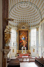 Frontale Farbfotografie eines Kirchenschiffs mit gold verzierter Bogendecke und einem hohen Gemälde hinter dem Altar. Das Zentrum des Gemäldes sind die Jungfrau Maria und das Christuskind, die von Menschen und Engeln umgeben sind. Über dem Gemälde sind Goldstrahlen und eine Krone zu sehen. Zwischen dem Gemälde und beiden Fenstern befinden sich helle Marmorsäulen. Auf dem Altar und an den Wänden sind goldene Figuren angebracht. Links vorne hängt an einem Pfeiler eine Marmorfigur mit goldenem Umhang und goldenem Heiligenschein.