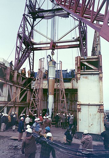 Nuclear device being lowered into its firing hole by a crane, with scientists in hard hats standing round
