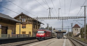 Kırmızı lokomotif treni platformdan geçirir; solda üçgen çatılı üç katlı bina var