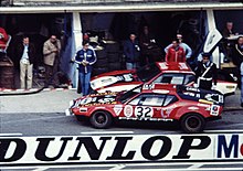 Photo d’époque d’une voiture de course rouge dans la ligne des stands du Mans