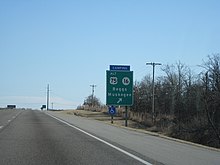 Et grønt skilt står "ALT US 75, SH 16, Beggs, Muskogee" med en pil.  En blå plak over den lyder "CAMPING".