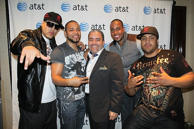 Five people. On the extreme left a man wearing a black denim pants, a jacket black synthetic leather, a white shirt, sunglasses and a black cap with red. Beside him a man wearing a blue denim pants and a gray shirt. In the center a man wearing gray suit and white shirt. On one side a man wearing a blue denim pants and a gray vest with coffee. To the far right a man wearing a black cap with red and black shirt with drawings and blue denim pants.