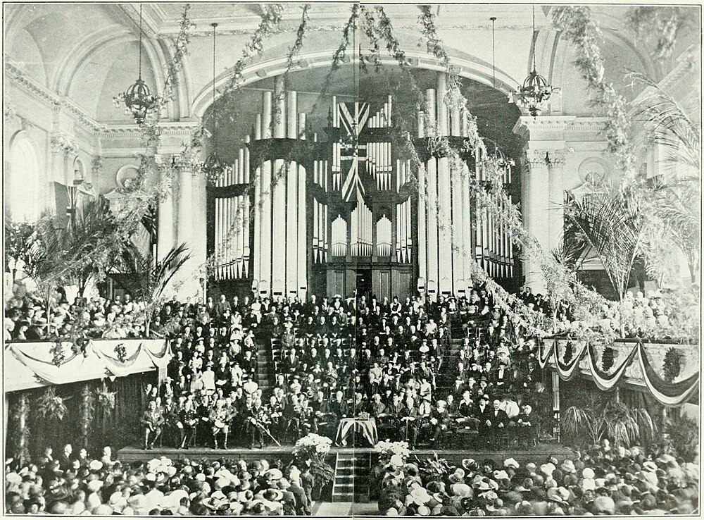 A grand hall with balconies on either side, organ pipes in the background with a stage in the front. People seated everywhere - on the balconies, on the hall floor, and facing the front on tiered seating.