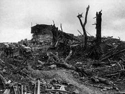 Heavily laden soldiers trudge through mud past the shattered remains of a concrete structure. Around them broken trees, steel beams and other pieces of debris have been strewn