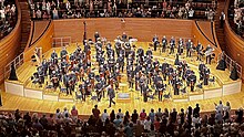 A symphony orchestra on a wooden stage receives a standing ovation from an audience.