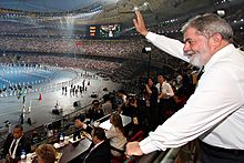 A man in the bleachers of a stadium waves during a parade. The venue seems crowded.