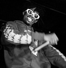 Black-and-white photo of a man in a hoodie with sunglasses, his hand holding the mic is moving and blurred.