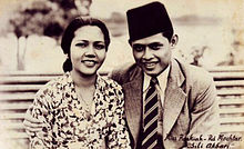 A promotional image showing a woman in a batik shirt sitting next to a man in a peci.