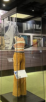 A yellow top and pants in a glass case in front of images of Taylor Swift from each album