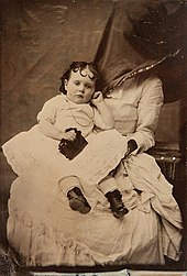 A toddler in a white lace dress frowning and clutching a black handbag, sat in the lap of a woman in a white lace dress, whose face is covered by loose black fabric.