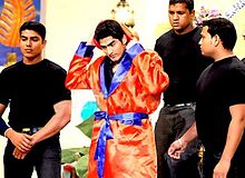 Four men standing. Middle of them is a young Indian male who wears a shiny orange dressing gown with blue border. His hands are behind his head. The other three men are all dressed in black and appear to be waiting around the man.