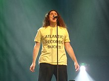 A man standing behind a microphone stand, wearing a yellow shirt that contains the text "Atlantic Records Sucks"