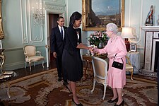 Michelle Obama and Elizabeth II shake hands and smile at each other as Barack enters the room in the background.