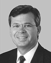 A man with dark hair and glasses wearing a black jacket and tie and white shirt