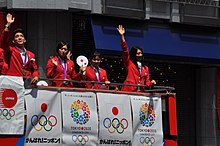 Ein Sportler und drei Sportlerinnen winken ins Publikum. Sie tragen rote Jacken und um den Hals ein lila Band, an dem die jeweilige Olympiamedaille baumelt. Der oben offene rote Bus ist mit Plakaten geschmückt, die für die Olympischen Spiele 2020 in Tokio werben. Gemäß Bildnamen entstand das Foto in Tokios Stadtteil Ginza.